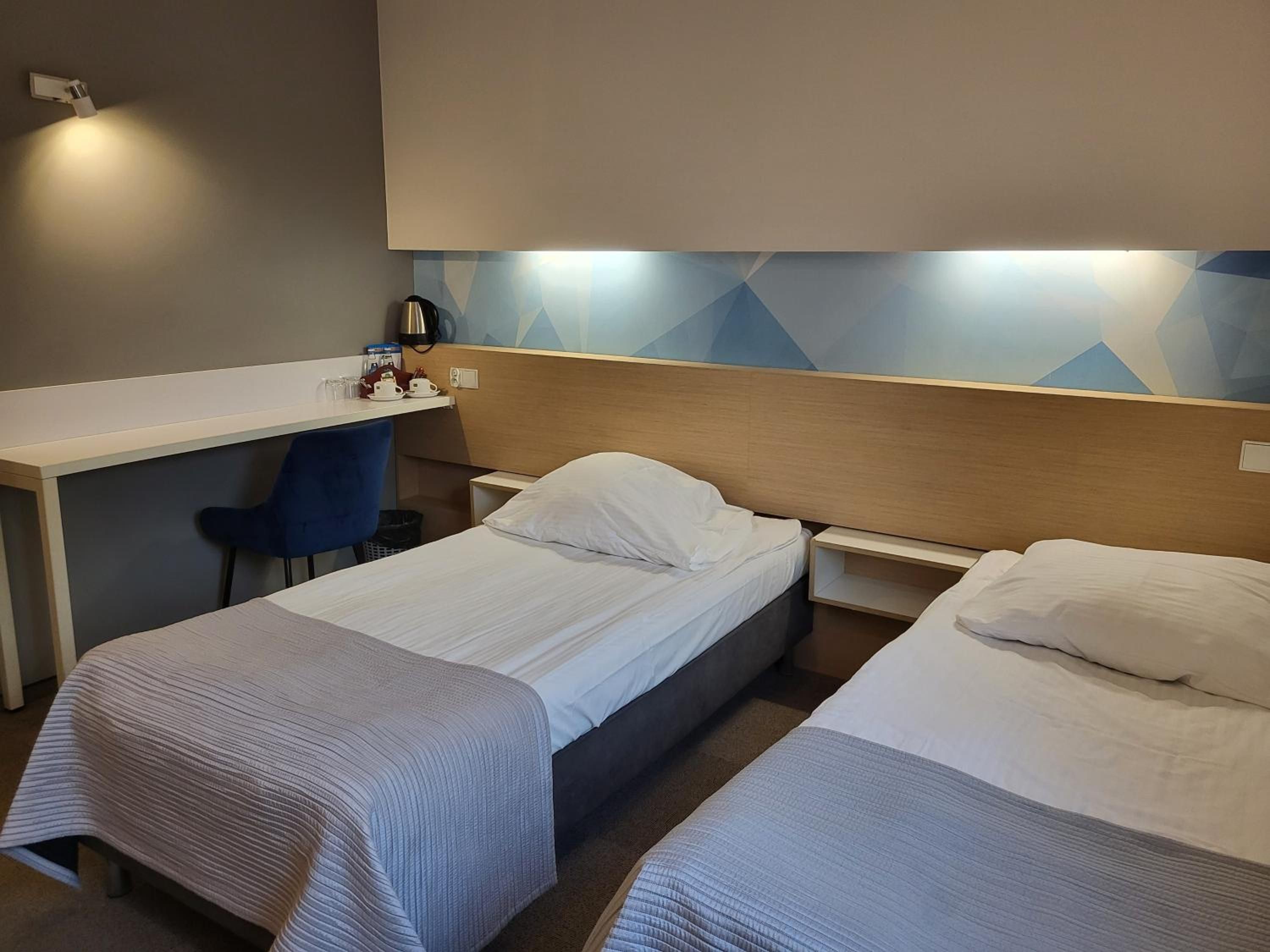 A hotel room with two single beds, a desk, a chair, and a wall-mounted light.