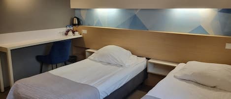 A hotel room with two single beds, a desk, a chair, and a wall-mounted light.