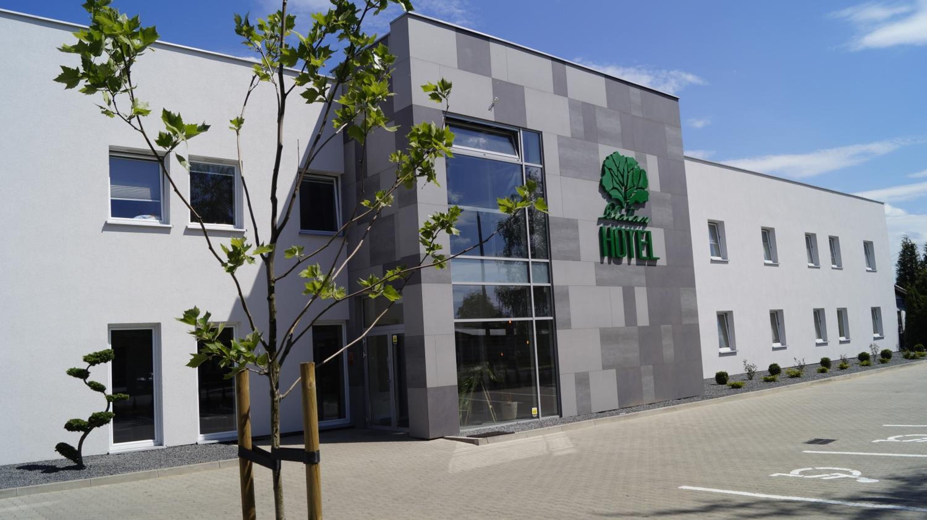 A modern hotel building with a large glass entrance, a logo with a tree, and a parking lot.