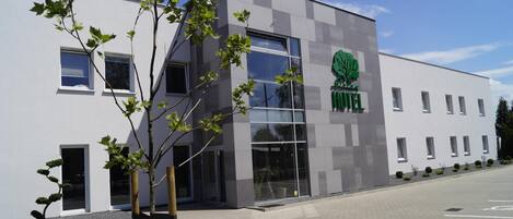 A modern hotel building with a large glass entrance, a logo with a tree, and a parking lot.