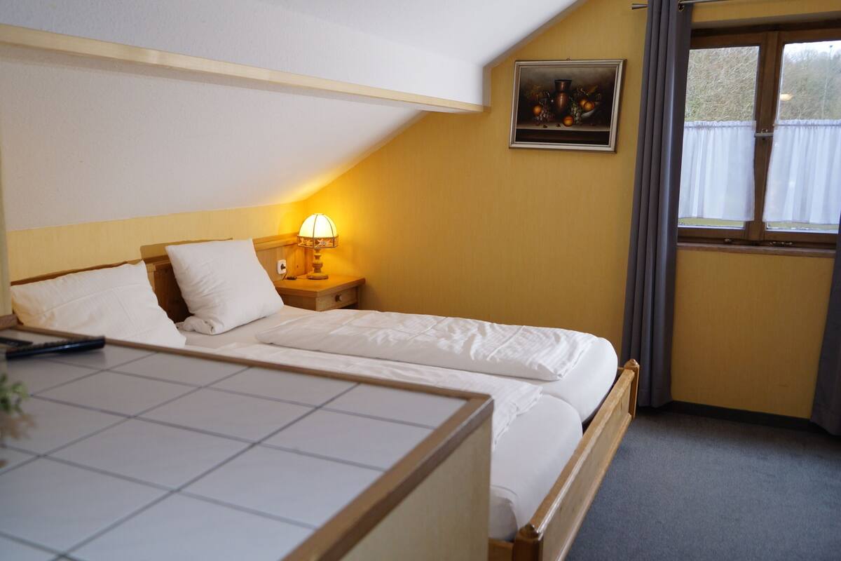 Habitación de hotel con cama, mesita de noche con lámpara, un cuadro en la pared y ventana con cortinas.