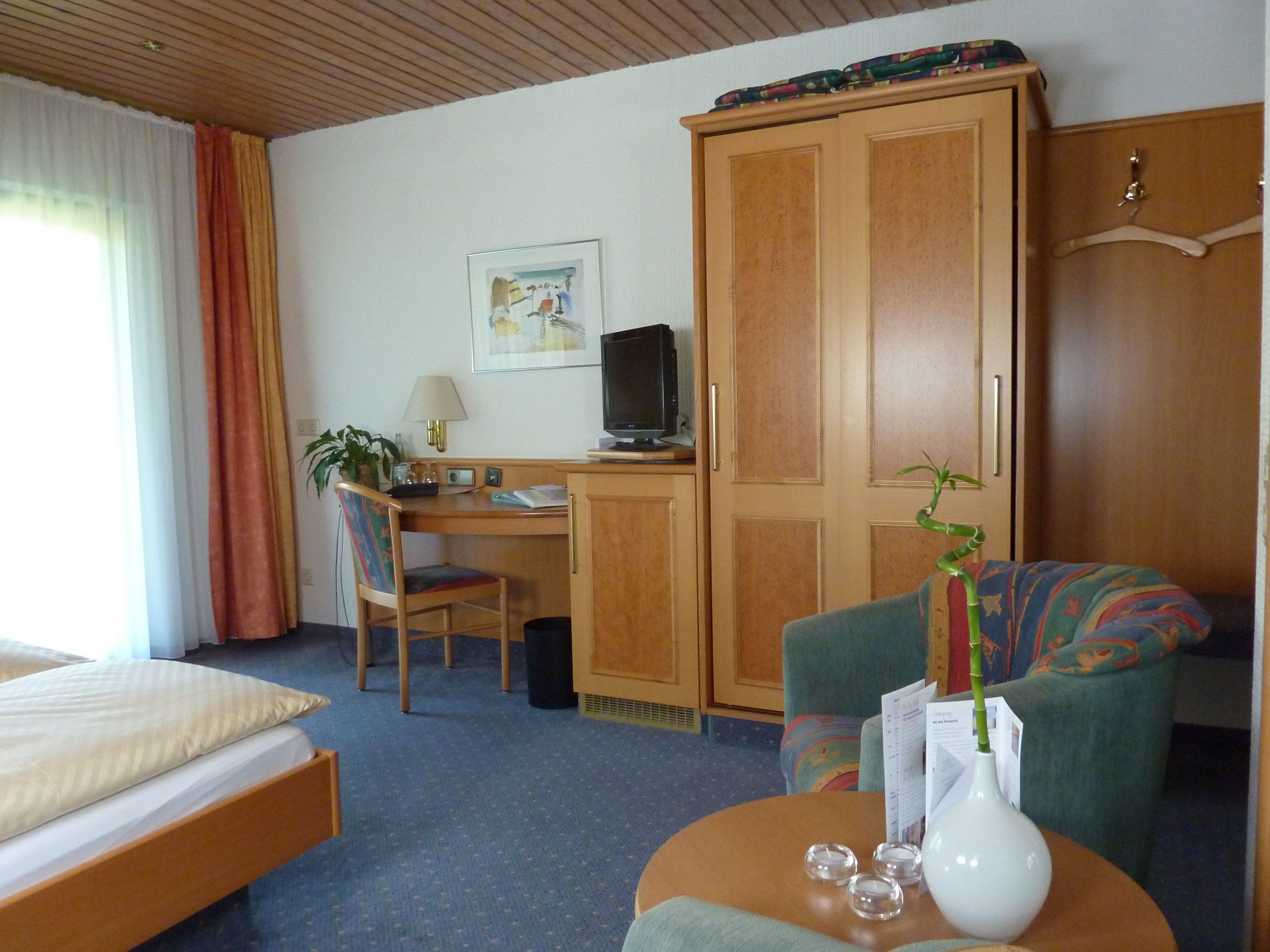 A hotel room with a bed, a desk with a computer, a chair, a wardrobe, and a small table with a vase and decorative items.