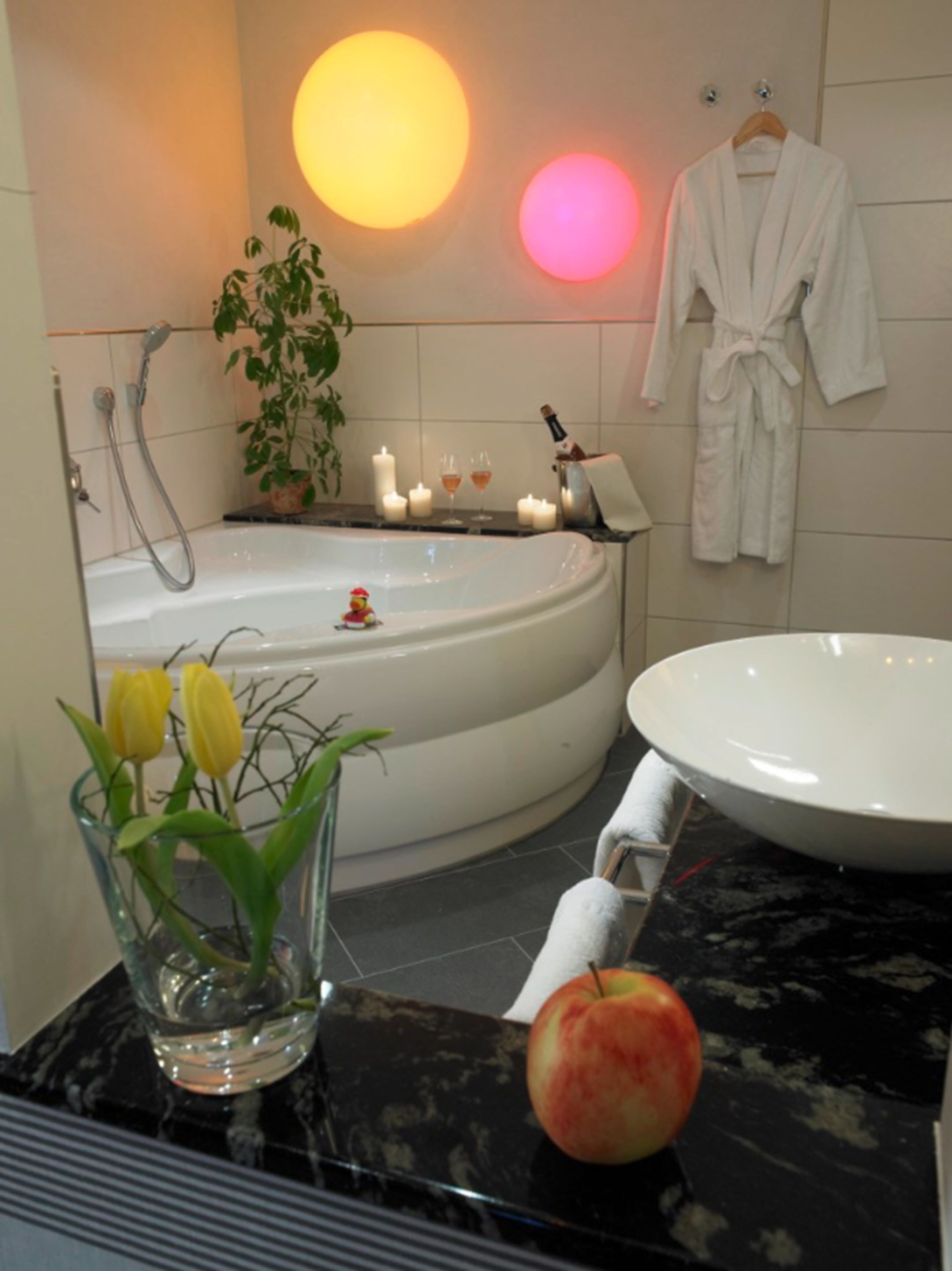 A modern bathroom with a freestanding bathtub, a round sink, a vase with yellow flowers, and a single apple on the counter.