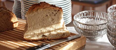 A loaf of bread on a wooden cutting board with a knife, surrounded by stacked plates and a metal strainer.