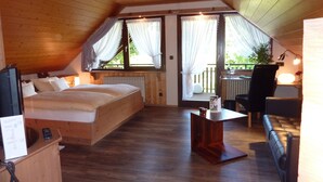 A cozy attic bedroom with a wooden bed, a small table, a leather sofa, and a view of the outdoors through large windows.