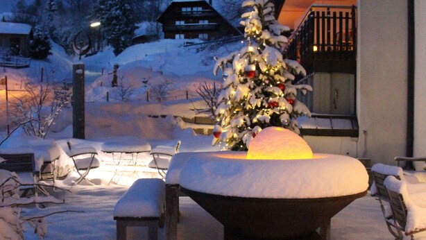 A snow-covered outdoor area with a snow-covered table and chairs, a Christmas tree, and a building with a balcony in the background.