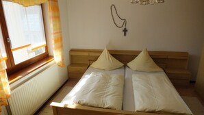 Una habitación pequeña y sencilla con una cama, un ventanal con cortinas, un radiador y una cruz en la pared.
