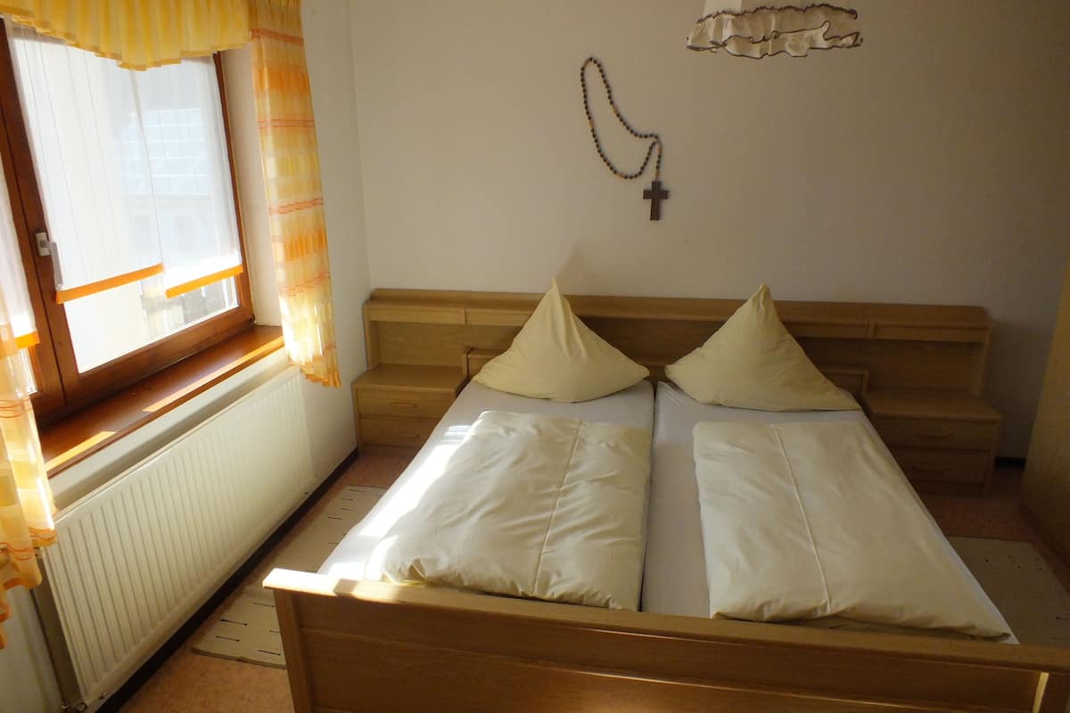 Une petite pièce simple, avec un lit, une fenêtre avec des rideaux, un radiateur et une croix accrochée au mur.