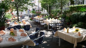An outdoor dining area with tables set for two, surrounded by greenery and potted plants.