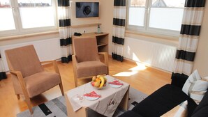 A room with two armchairs, a small table with a fruit bowl, a black sofa, and a TV mounted on the wall. - Hotel Anika (Neuenburg am Rhein)
