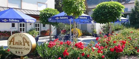 Un letrero de hotel con logotipo de restaurante-café, mesas al aire libre con sombrillas azules y flores rojas.