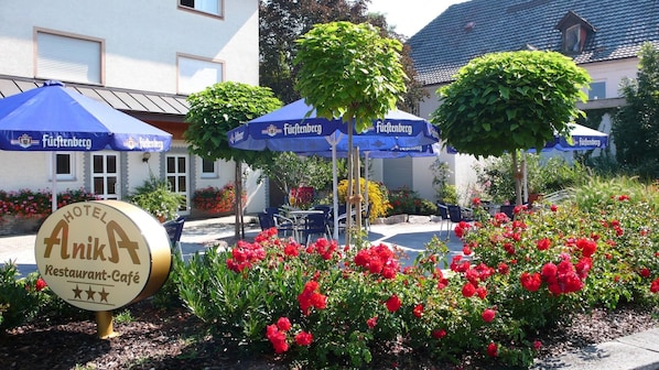 A hotel sign with a restaurant-café logo, outdoor seating with blue umbrellas, and red flowers.