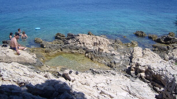 Una costa rocosa con agua azul cristalina y algunas personas nadando.
