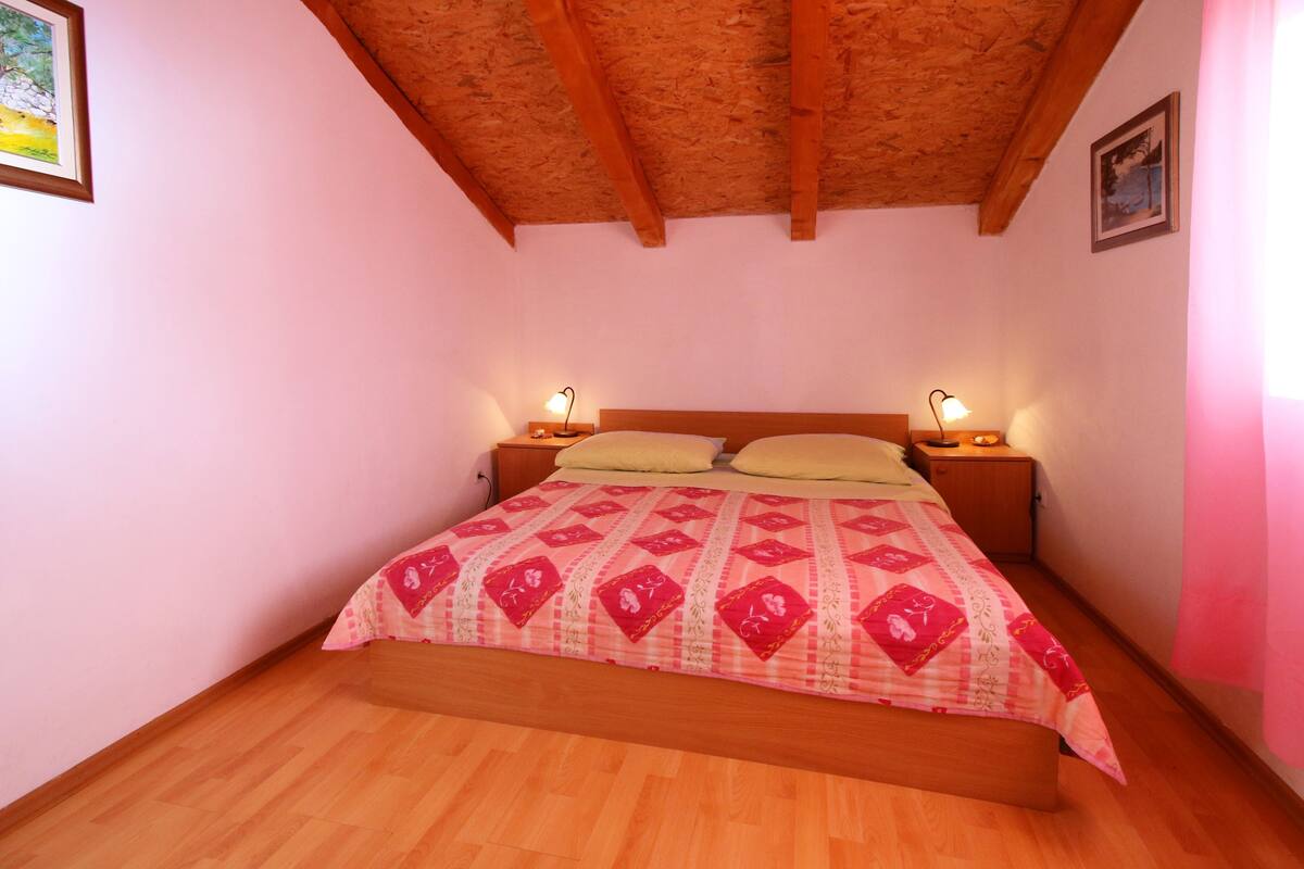 A bedroom with a wooden ceiling, a bed with a patterned bedspread, two bedside lamps, and a framed picture on the wall.