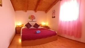 Un dormitorio con techo de madera, una cama con una colcha roja, dos mesitas de noche y una ventana con cortinas rosadas.