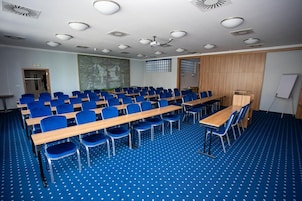 Un salón de conferencias con hileras de sillas azules, mesas de madera y una pizarra.