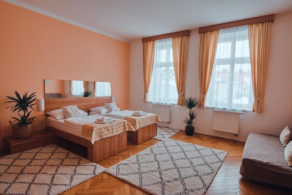 A bedroom with two beds, a wooden headboard, a large window with curtains, a potted plant, and a sofa.