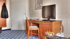 A hotel room with a wooden desk, a chair, a television, a small table with a fruit bowl, and a water bottle.