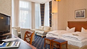 A hotel room with a bed, a desk with a TV, a chair, a sofa, and a large window with curtains.