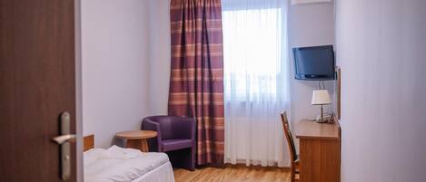 A hotel room with a bed, a desk with a lamp, a chair, a television, and a window with curtains.