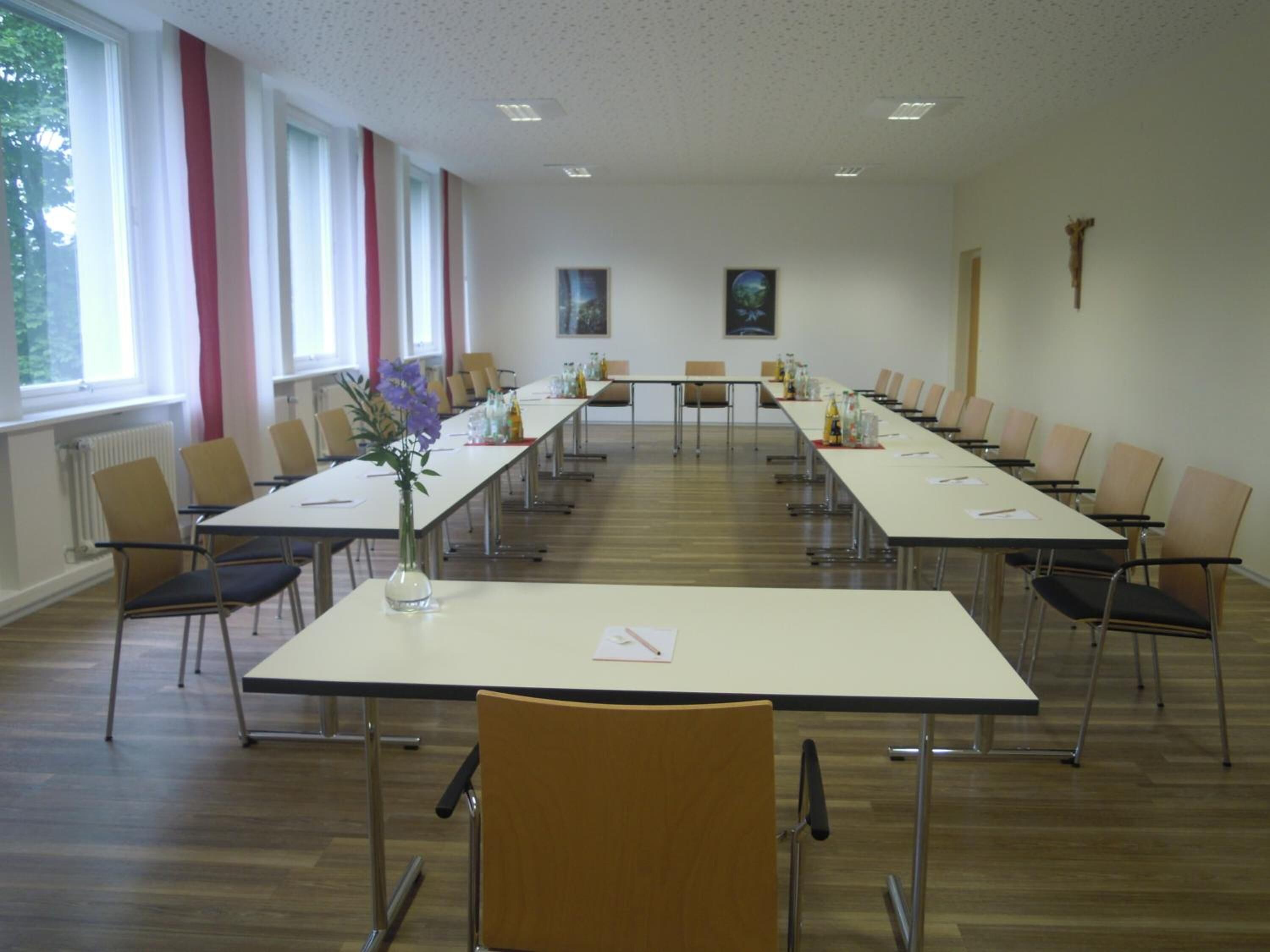 Ein Konferenzraum mit langen Tischen und Stühlen, großen Fenstern und einem Kruzifix an der Wand.