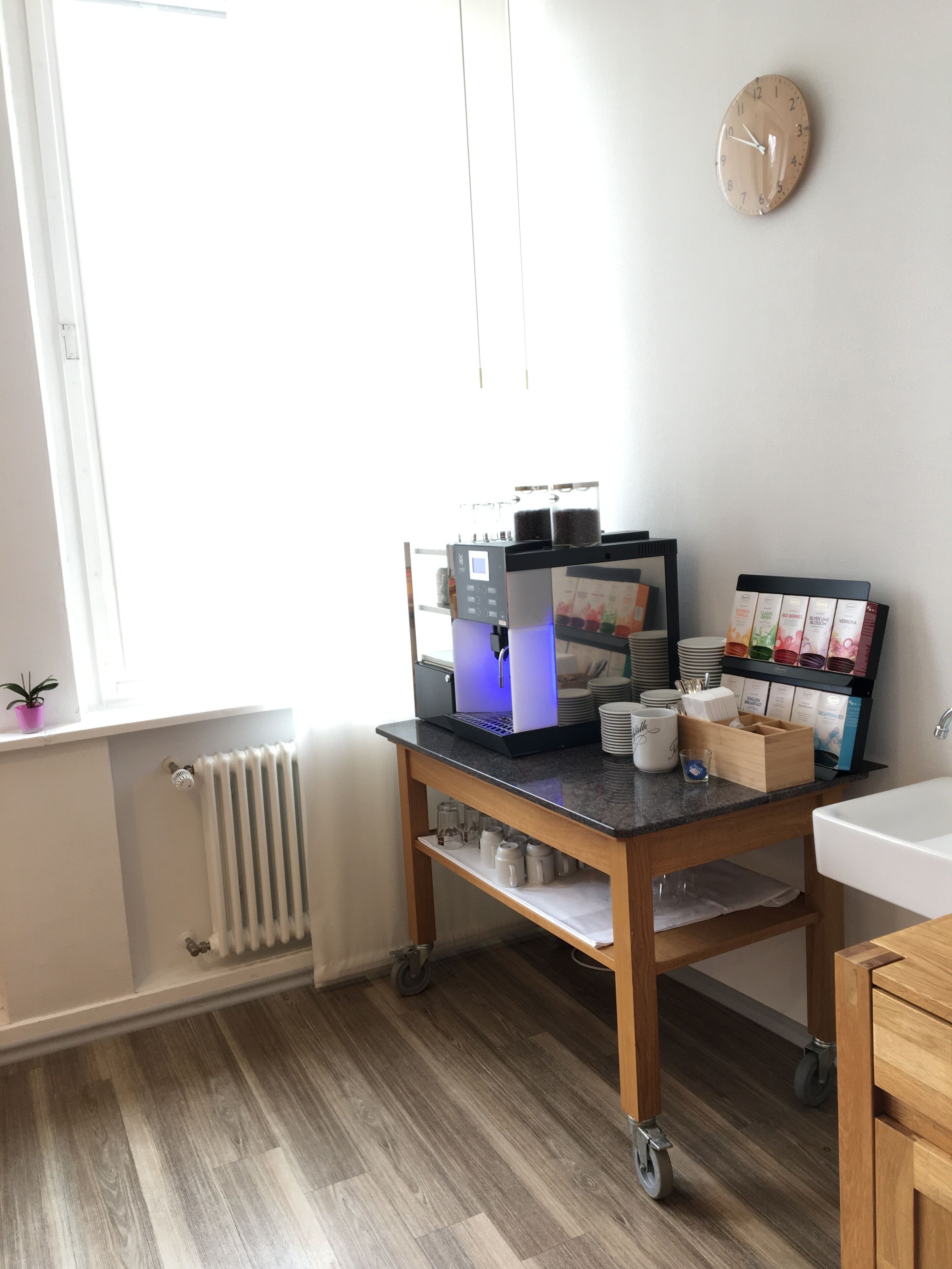 A small, well-lit room with a wooden floor, a desk with a laptop, a coffee machine, and a clock on the wall.