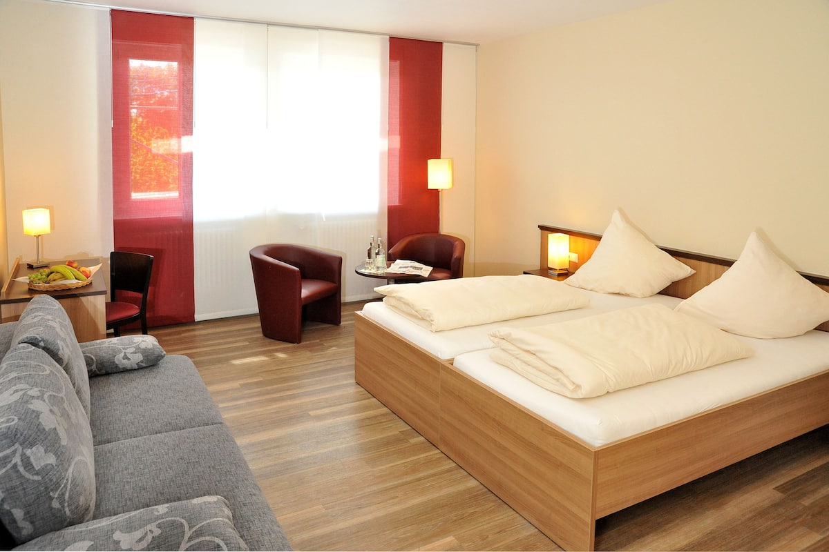 A hotel room with a bed, a sofa, a chair, a small table with a lamp, and a window with curtains.