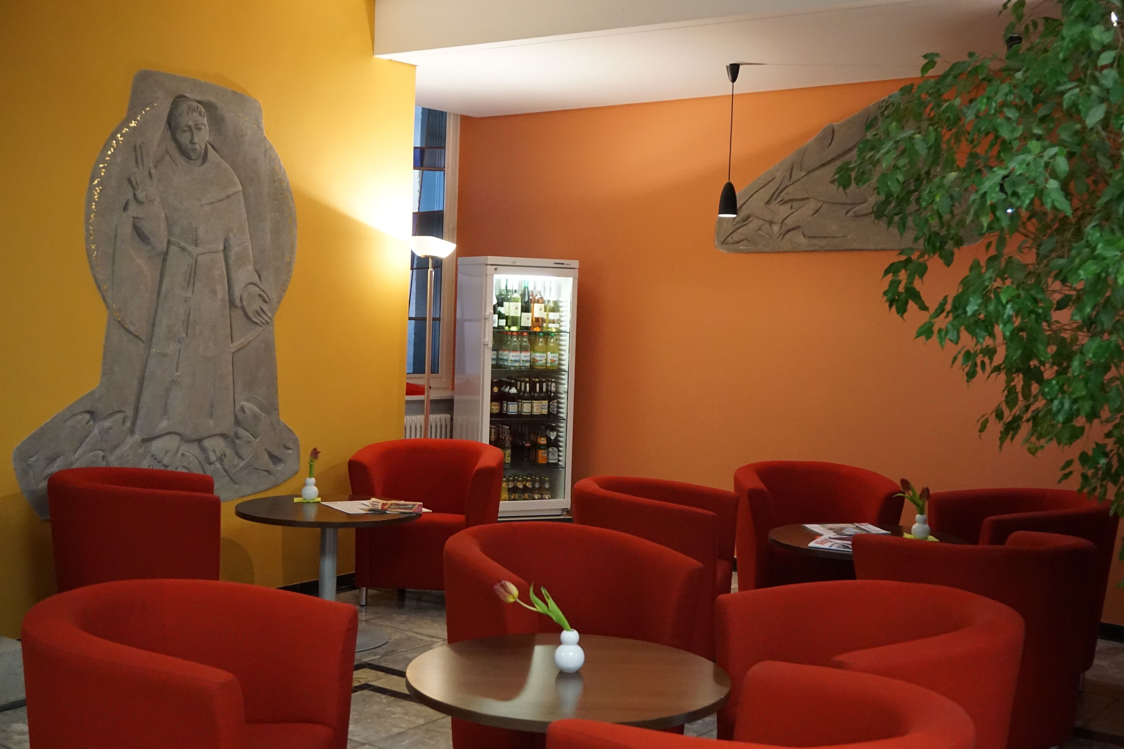 A restaurant with red chairs, a large sculpture on the wall, and a refrigerator with bottles.