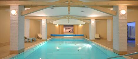 Una piscina interior con vista despejada al área de la piscina, sillas de descanso y una puerta que conduce a otra habitación.