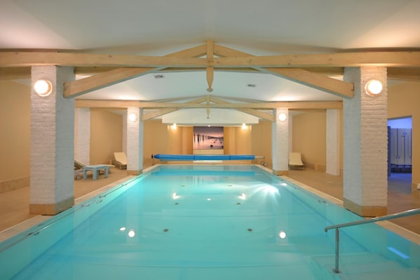 Una piscina interior con vista despejada al área de la piscina, sillas de descanso y una puerta que conduce a otra habitación.