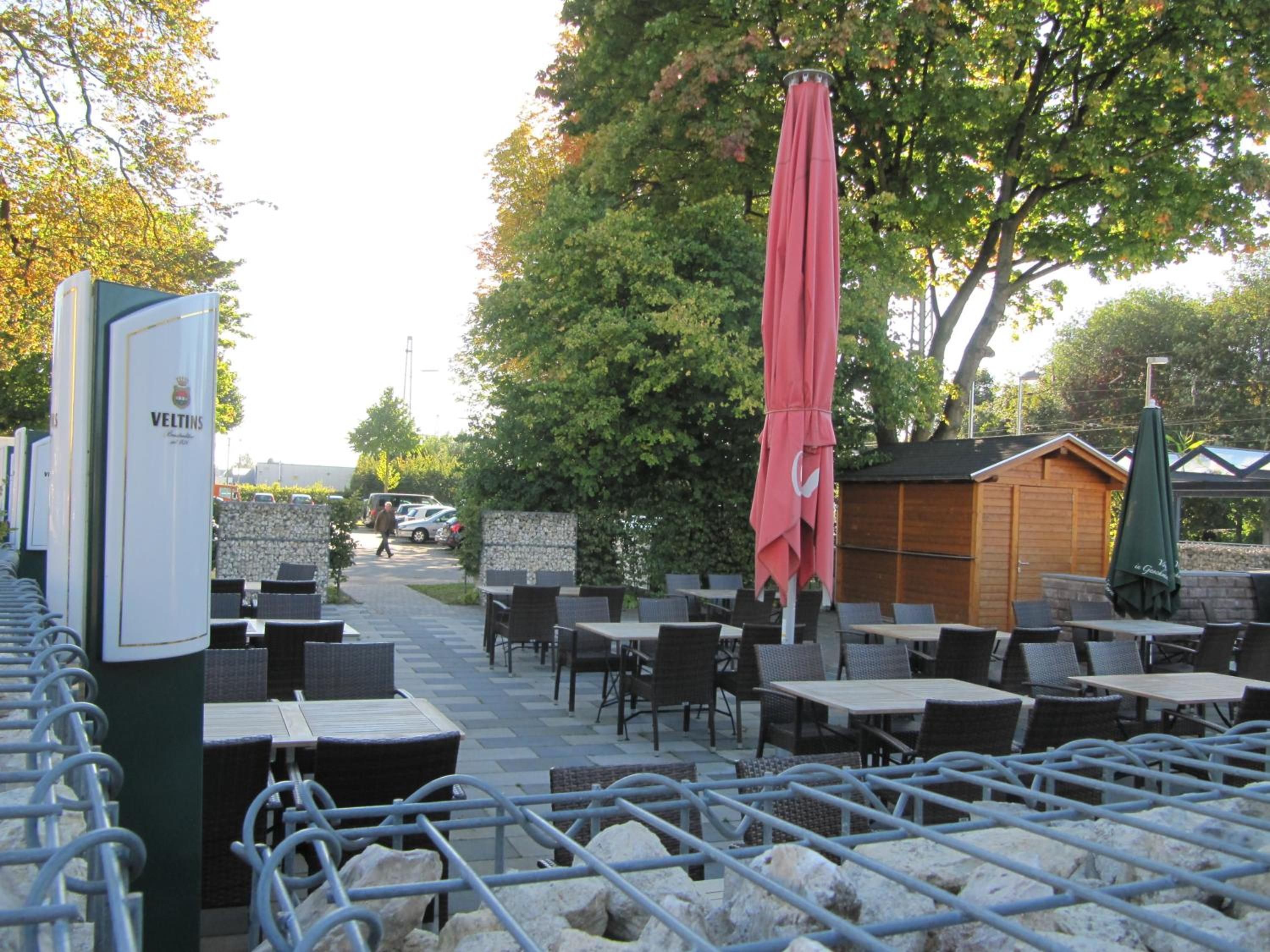An outdoor seating area with tables and chairs, a red umbrella, and a sign for Veltins.