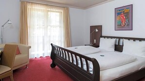 Habitación de hotel con cama, una silla, escritorio, lámpara y ventana con cortinas.