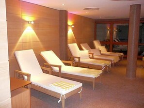 A row of sun loungers with cushions, a wooden floor, and wall-mounted lights.