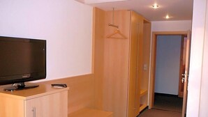 A hotel room with a TV on a stand, a wooden wardrobe, a bench, and a door leading to another room. - Landhotel Grv Nenbach (Bad Groenenbach)
