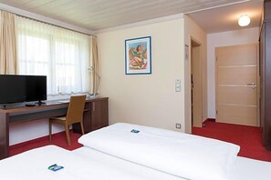 A hotel room with a bed, a desk with a television, a chair, a painting on the wall, and a door.