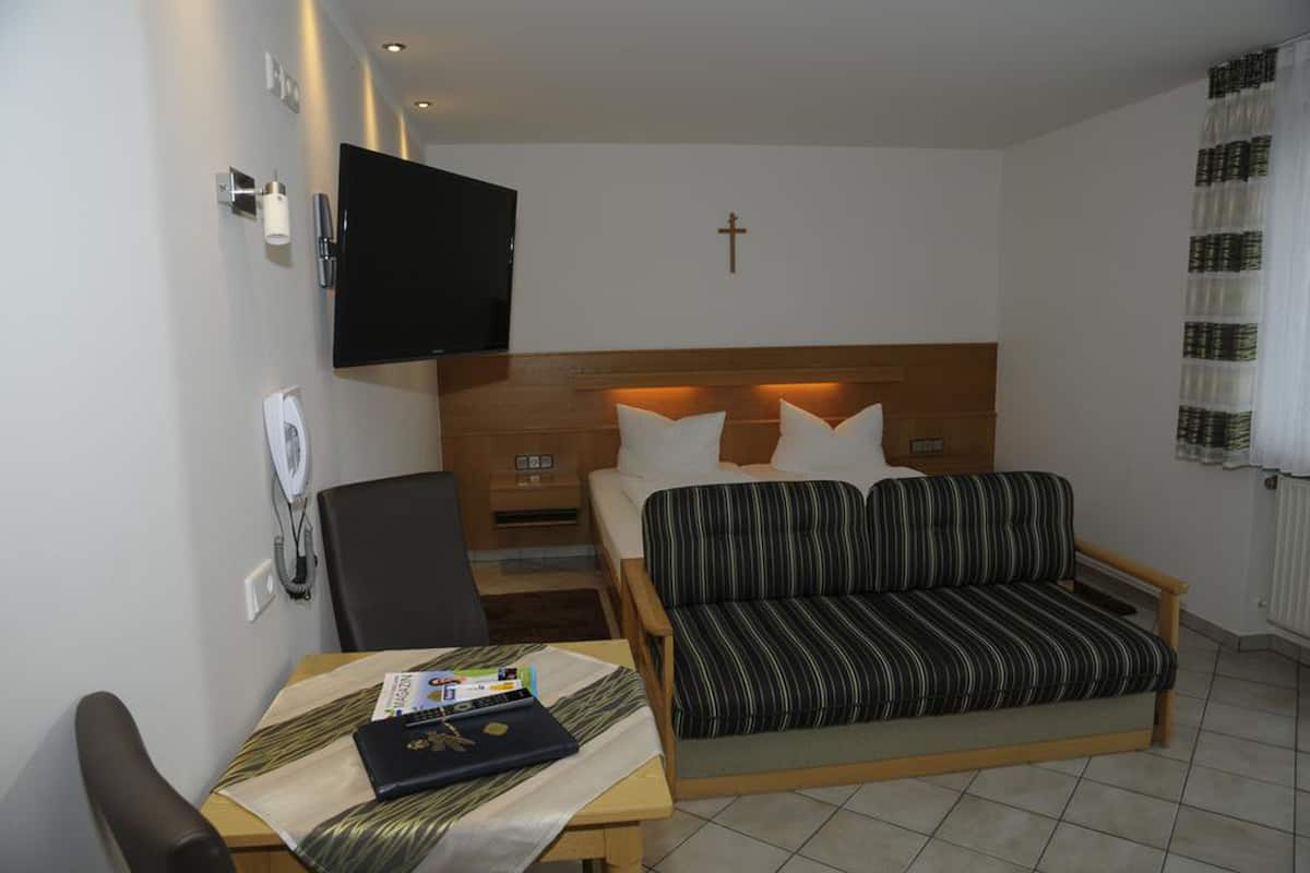 Una habitación de hotel moderna con una cama, un televisor, una cruz en la pared y una pequeña mesa con una computadora portátil y un libro.