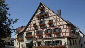 Il s’agit d’un bâtiment traditionnel à colombages, doté de nombreuses fenêtres, dont certaines sont ornées de jardinières, et d’une enseigne indiquant qu’il s’agit d’un restaurant.
