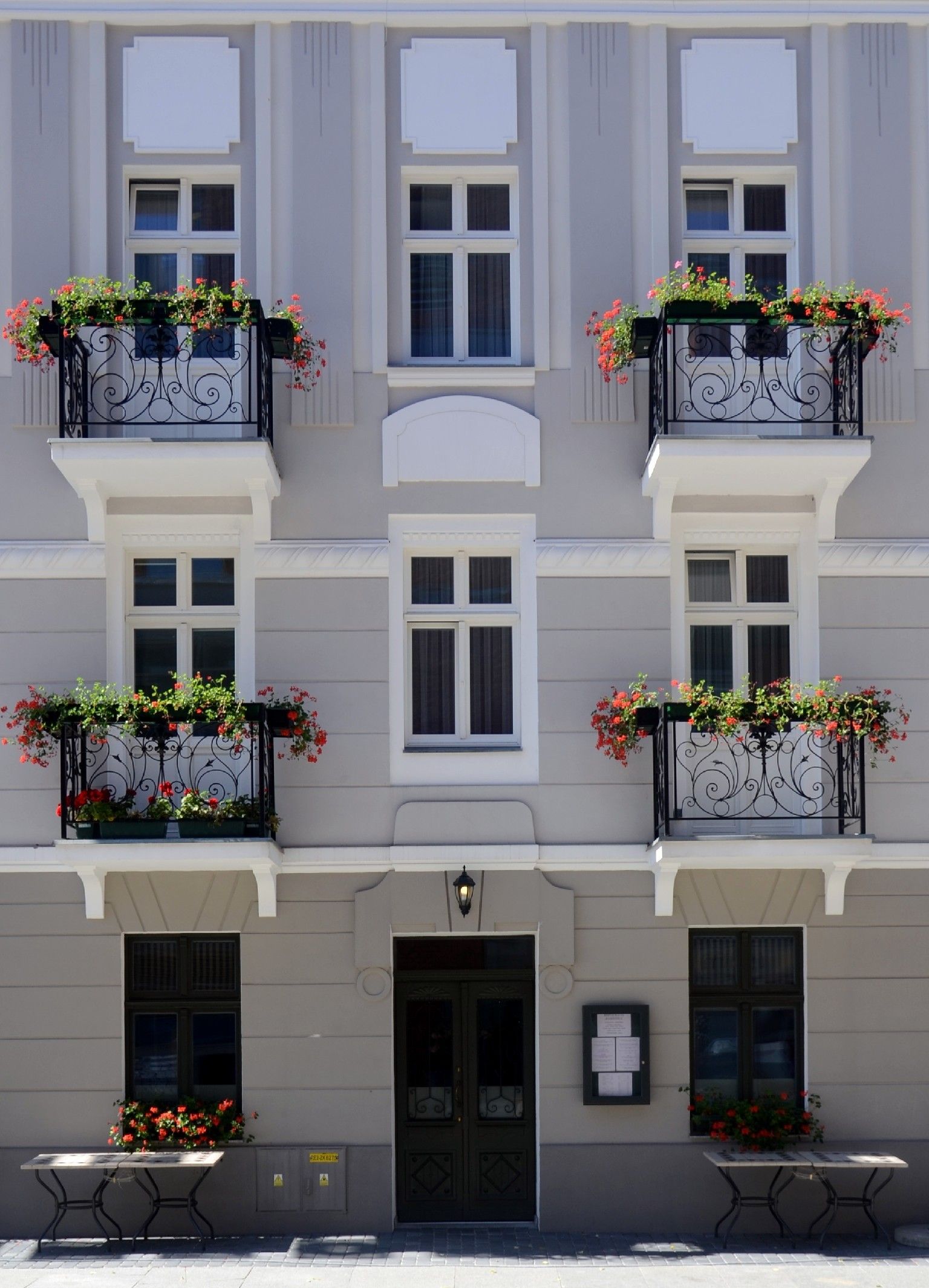 Ein mehrstöckiges Gebäude mit Balkonen, die mit Blumenkästen geschmückt sind, einem zentralen Eingang mit einer Tür und einem kleinen Tisch mit Papieren im Erdgeschoss.