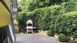 A paved courtyard with a stone barbecue grill, surrounded by dense greenery and a stone wall. - Apartments Veno (Lasko)