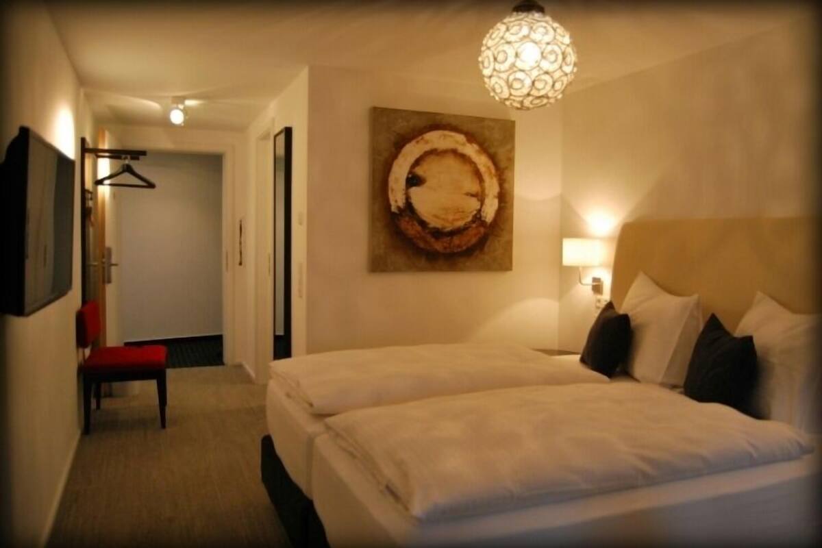 A hotel room with two beds, a hanging light fixture, a painting on the wall, and a chair in the corner.