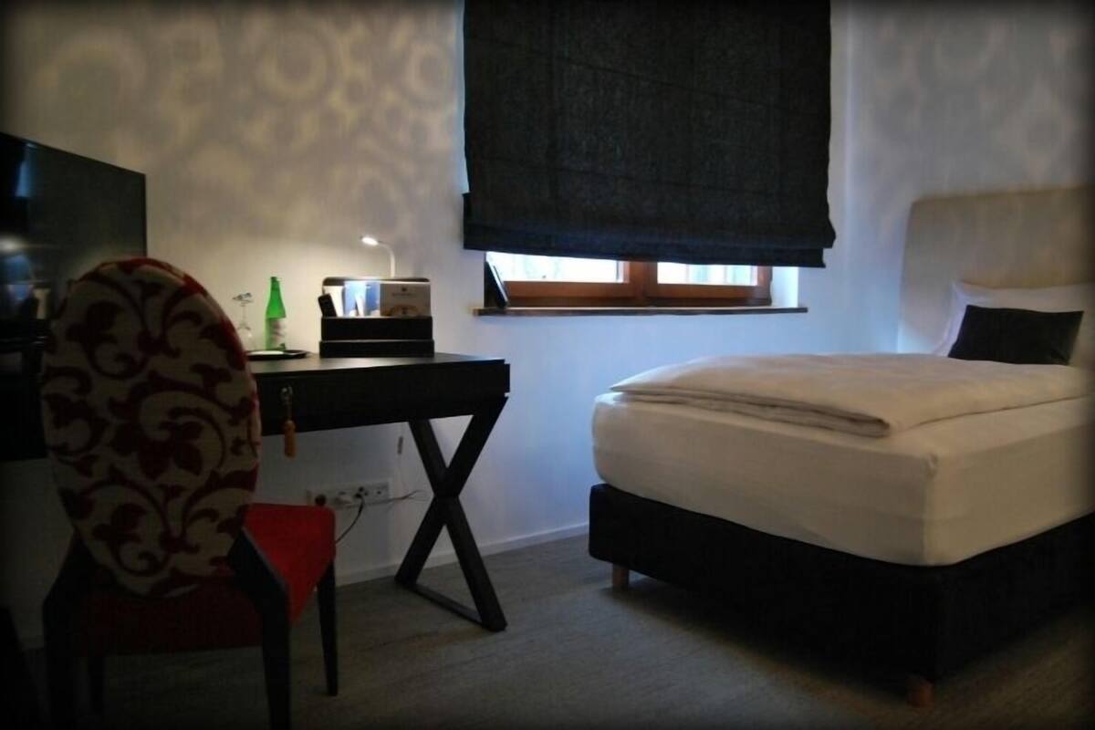 A bedroom with a bed, a desk, a chair, and a window with a blind.