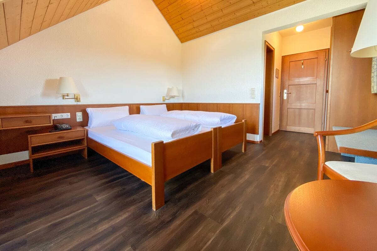 A bedroom with two beds, wooden headboards, bedside tables with lamps, a wooden chair, and a door.