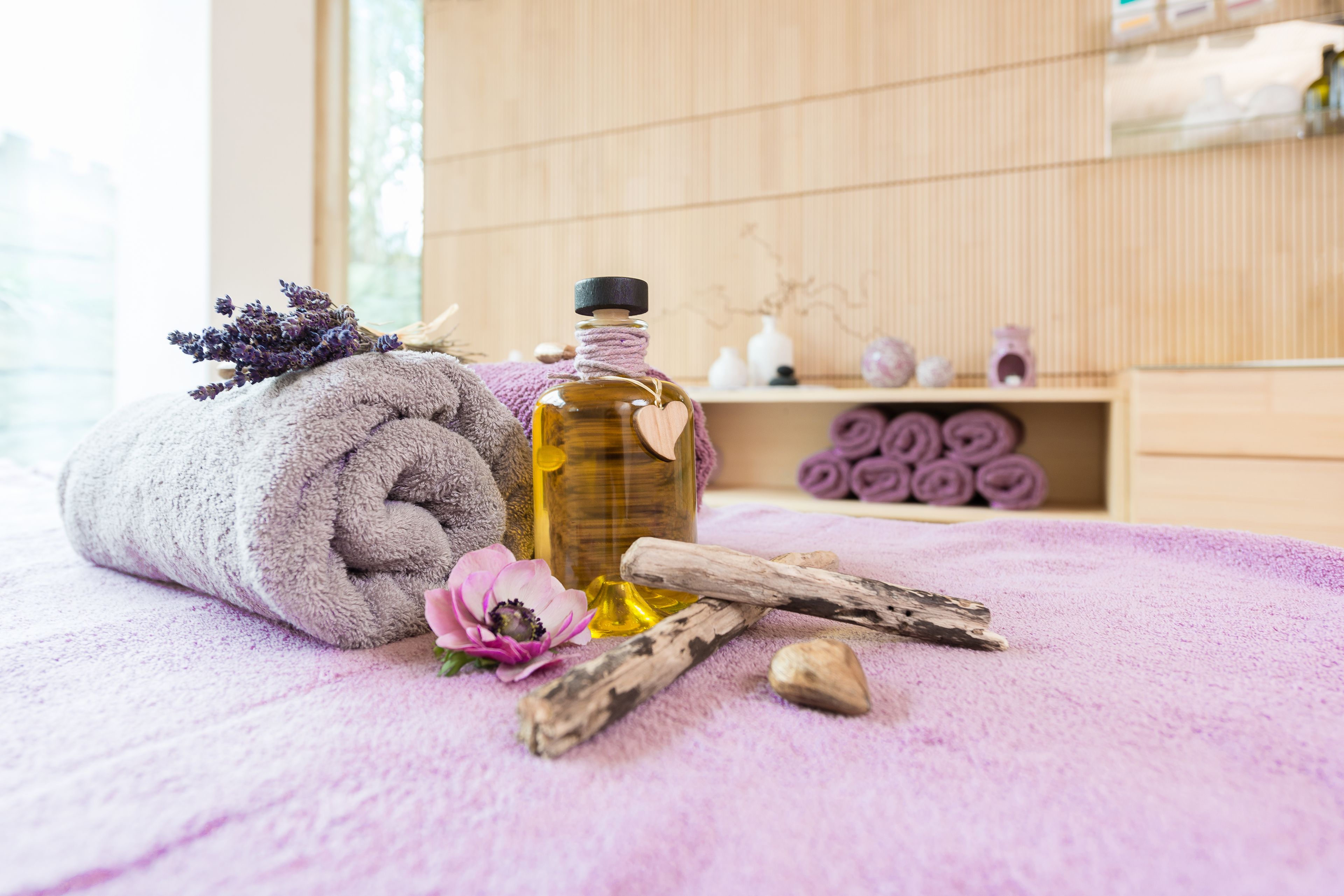 A neatly rolled towel, a bottle of oil, a flower, and wooden sticks on a purple towel in a room with wooden walls and shelves.