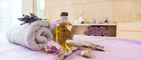 A neatly rolled towel, a bottle of oil, a flower, and wooden sticks on a purple towel in a room with wooden walls and shelves.