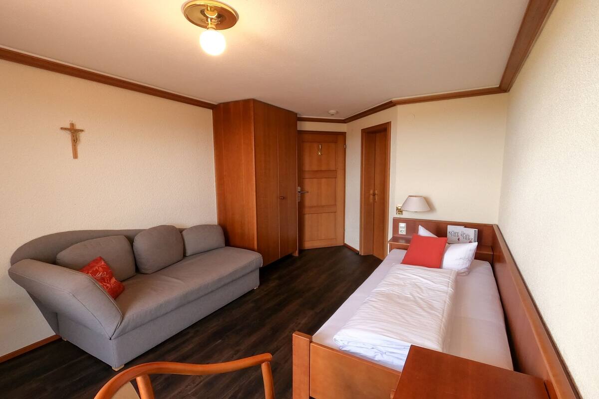 A room with a bed, a sofa, a wooden chair, a cross on the wall, and a door.