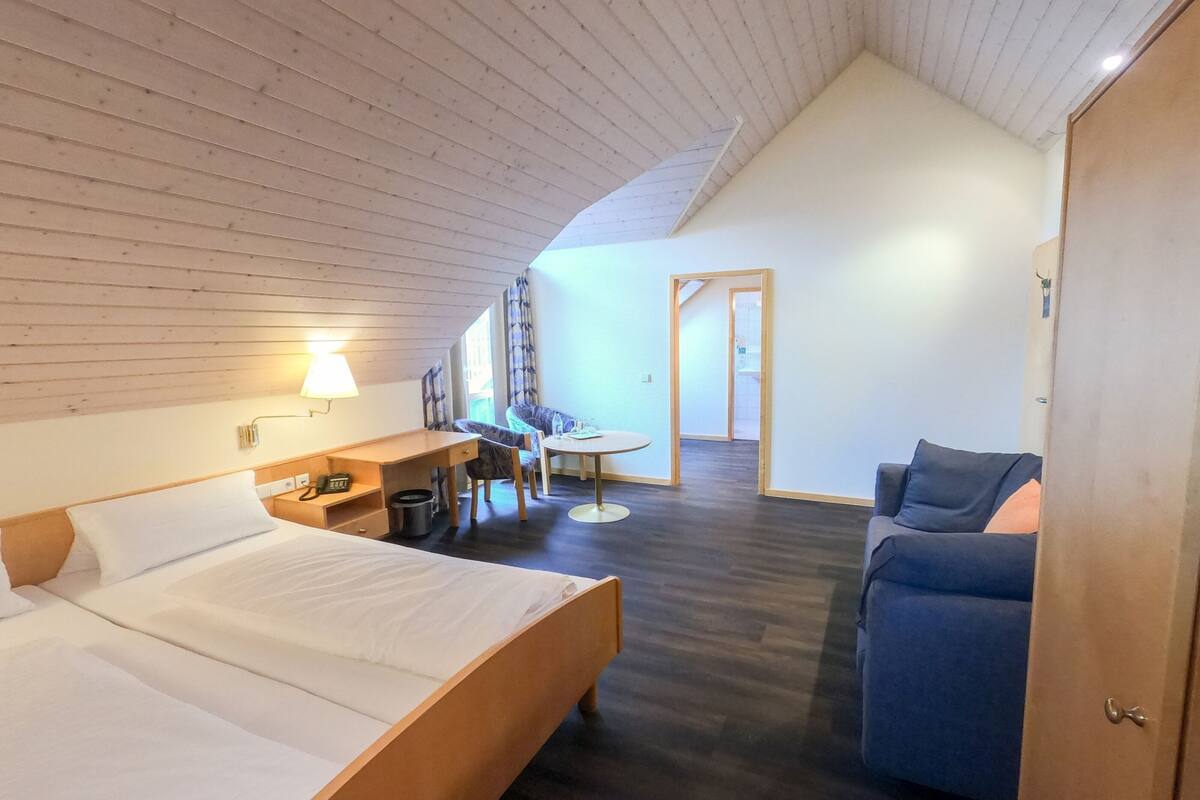 A hotel room with a wooden ceiling, a bed, a desk with a chair, a sofa, and a small table.