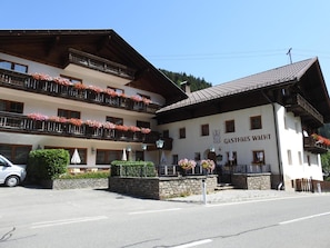 A multi-story building with balconies adorned with flowers, a sign reading 'GASTHAUS WACHT,' and a stone wall in the foreground.
