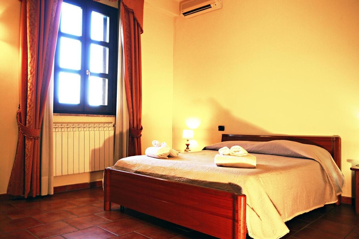 A hotel room with a bed, a window with curtains, an air conditioning unit, a radiator, and a bedside lamp.