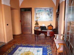 A hallway with a wooden door, a desk with a laptop, a chair, and a tiled floor with a decorative border.