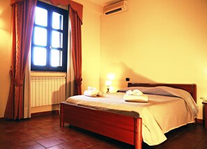 A hotel room with a bed, a window with curtains, an air conditioning unit, a radiator, and a bedside lamp.